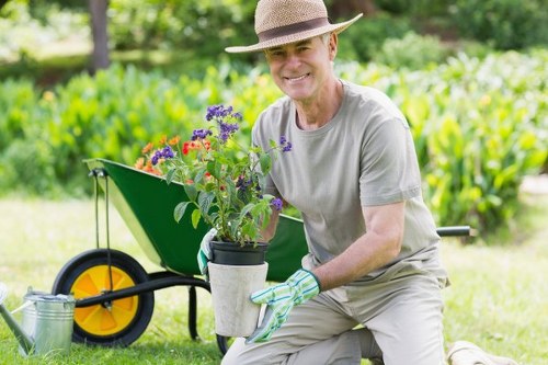 A gardener wearing a straw hat, light grey t-shirt, and gardening gloves is kneeling on a well-maintained lawn in a lush outdoor garden in Chelsea. They are holding a black pot filled with vibrant purple and red flowering plants, with green foliage visible. Behind the gardener, there is a bright green wheelbarrow with large yellow and black wheels, positioned on the grass, which appears to be dense and neatly trimmed. The background features a variety of trees and shrubs, contributing to the lush, natural environment of the landscaped outdoor space. The scene is illuminated by natural daylight, suggesting a sunny or partly cloudy day, emphasizing the healthy, vibrant colors of the plants and the well-kept garden layout. This setting exemplifies professional gardening and lawn care services offered by Garden Maintenance Chelsea, highlighting attention to detail and a quality outdoor environment typical of Chelsea gardens.