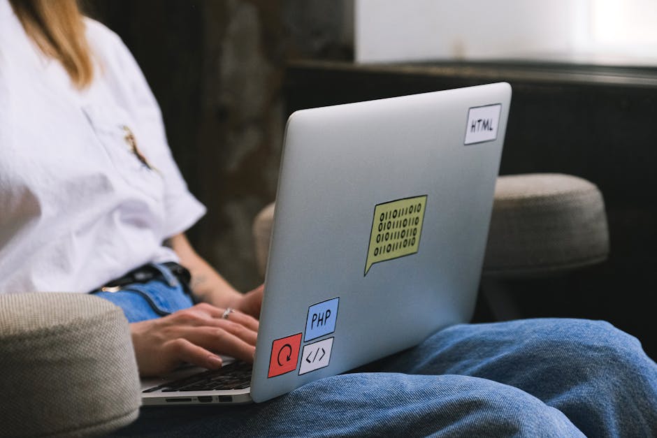 A black and white photograph of a workspace with a laptop displaying lines of code on its screen, positioned on a dark wooden desk against a textured brick wall background. To the right of the laptop,